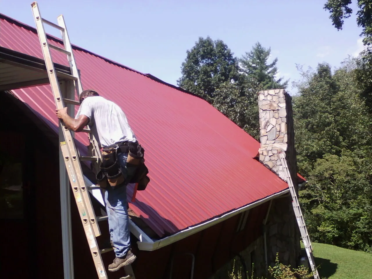 Roofer on a ladder beside a red metal roof and stone chimney — Soffit & Fascia Repair by Roofing Brothers in West Townsend
